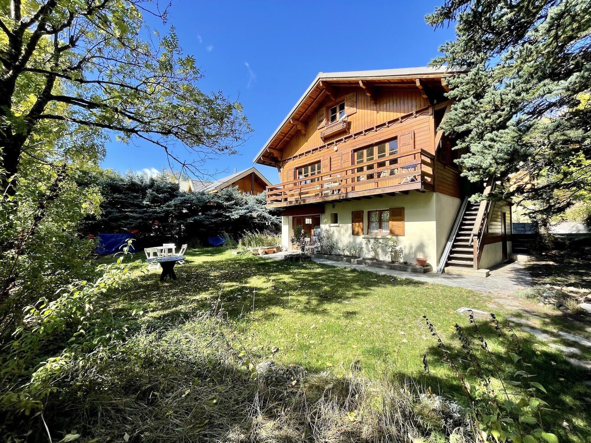 A wooden chalet with a balcony, surrounded by greenery and a clear sky.
