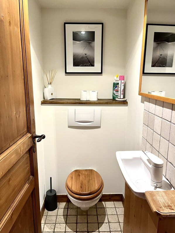 A bathroom with a wooden door, a toilet with a wooden seat, a white sink, a mirror, and two framed pictures on the wall.
