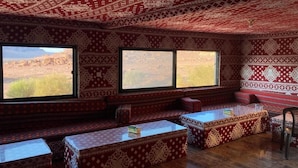Una habitación con paredes decoradas, amplias ventanas, suelo de madera y una zona de estar con bancos y mesas.