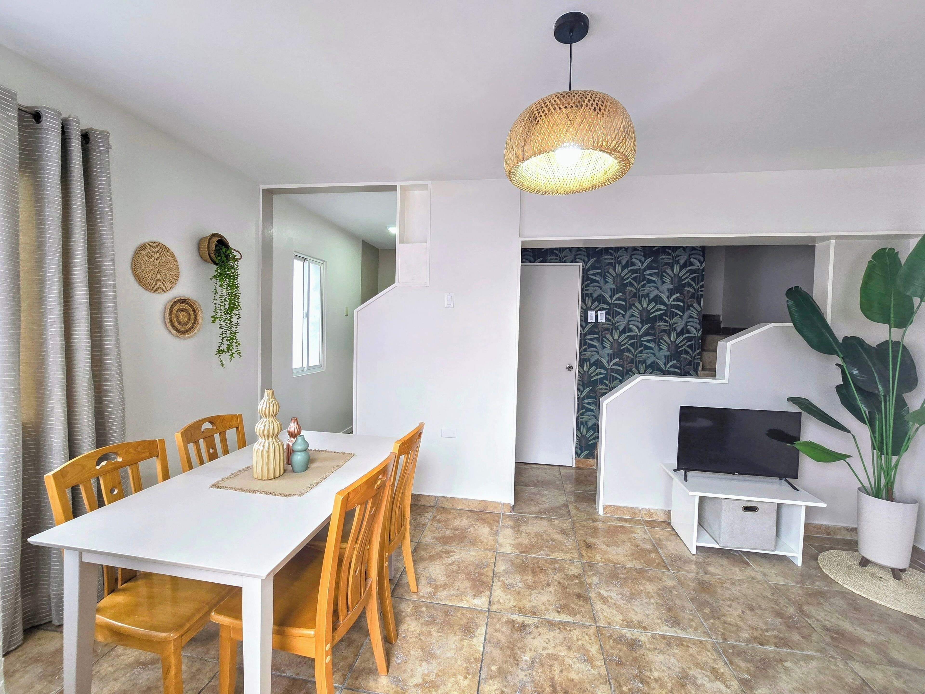 A dining area with a white table and wooden chairs, a hanging woven light fixture, and a view into a kitchen area with a TV and potted plants.