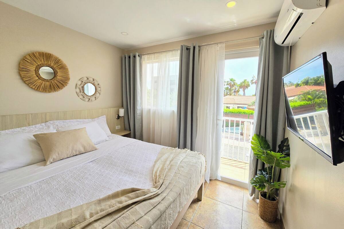 A hotel room with a bed, a TV, a wall-mounted air conditioner, and a view of the outside balcony.