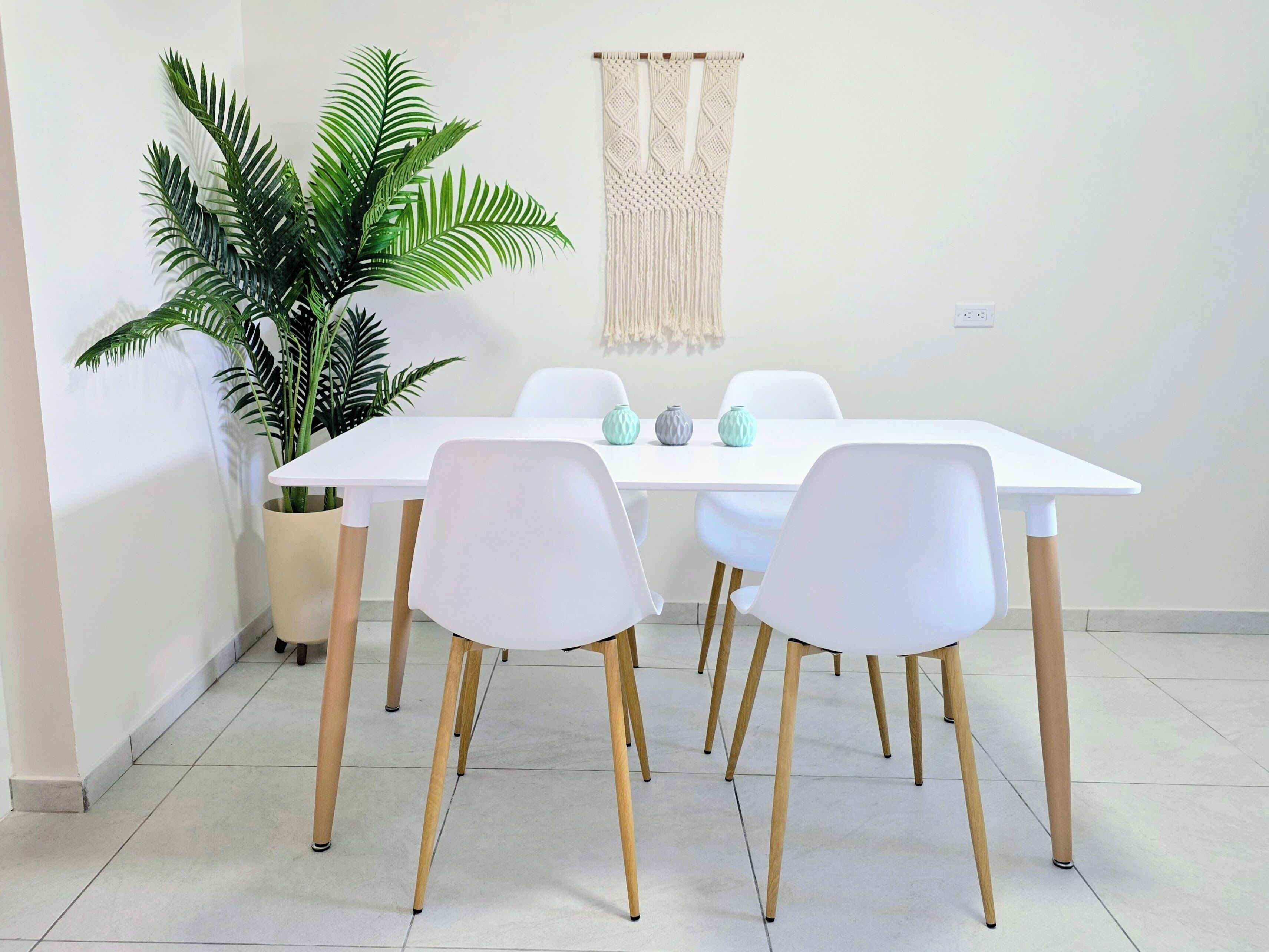 A dining area with a white table, four white chairs with wooden legs, a large green plant, and a macrame wall hanging.