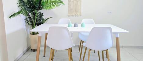 A dining area with a white table, four white chairs with wooden legs, a large green plant, and a macrame wall hanging.