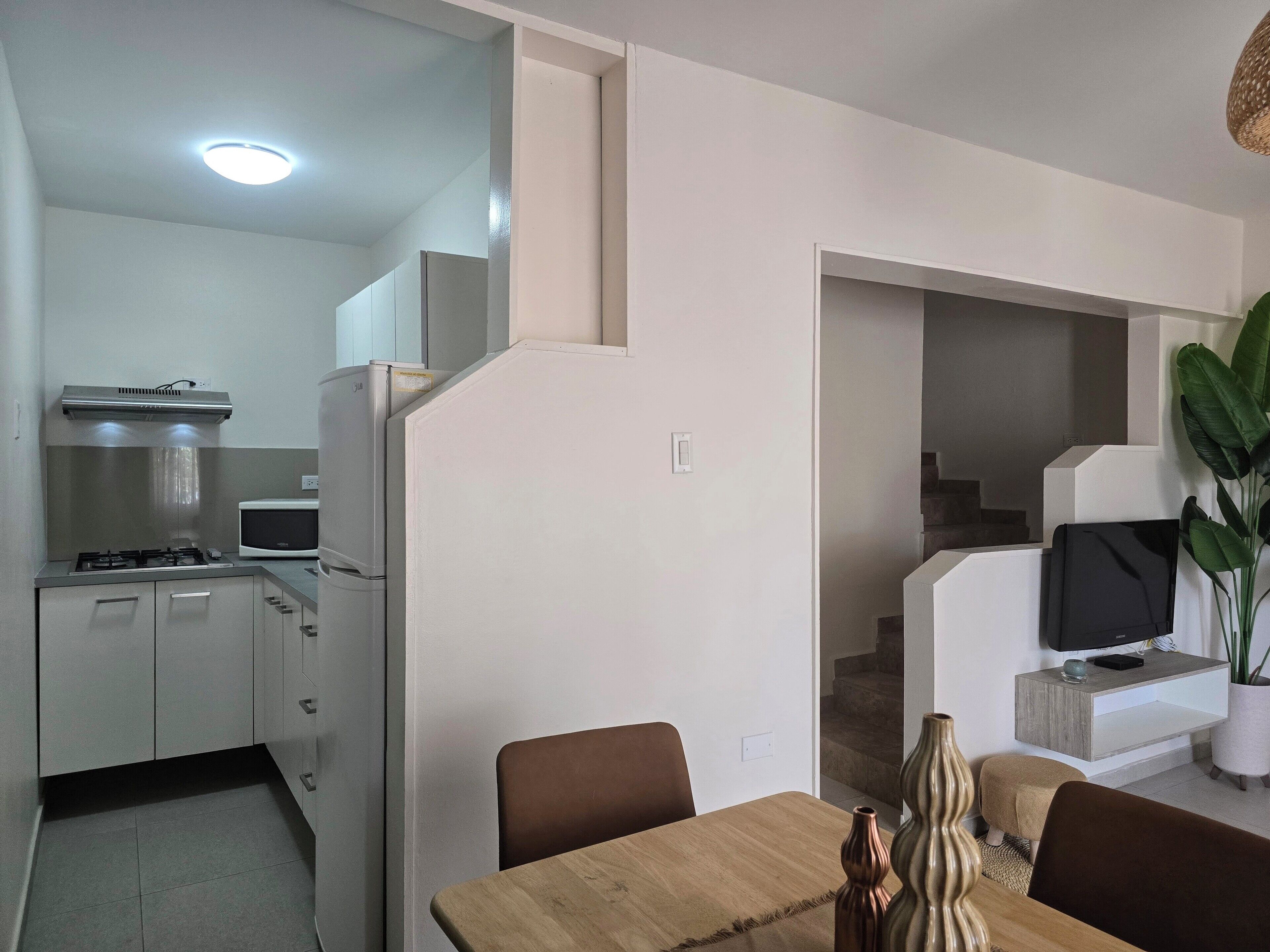 Una cocina moderna con estufa a gas, microondas y horno empotrado. Hay un comedor con mesa y sillas de madera, y un televisor montado en una pared blanca.