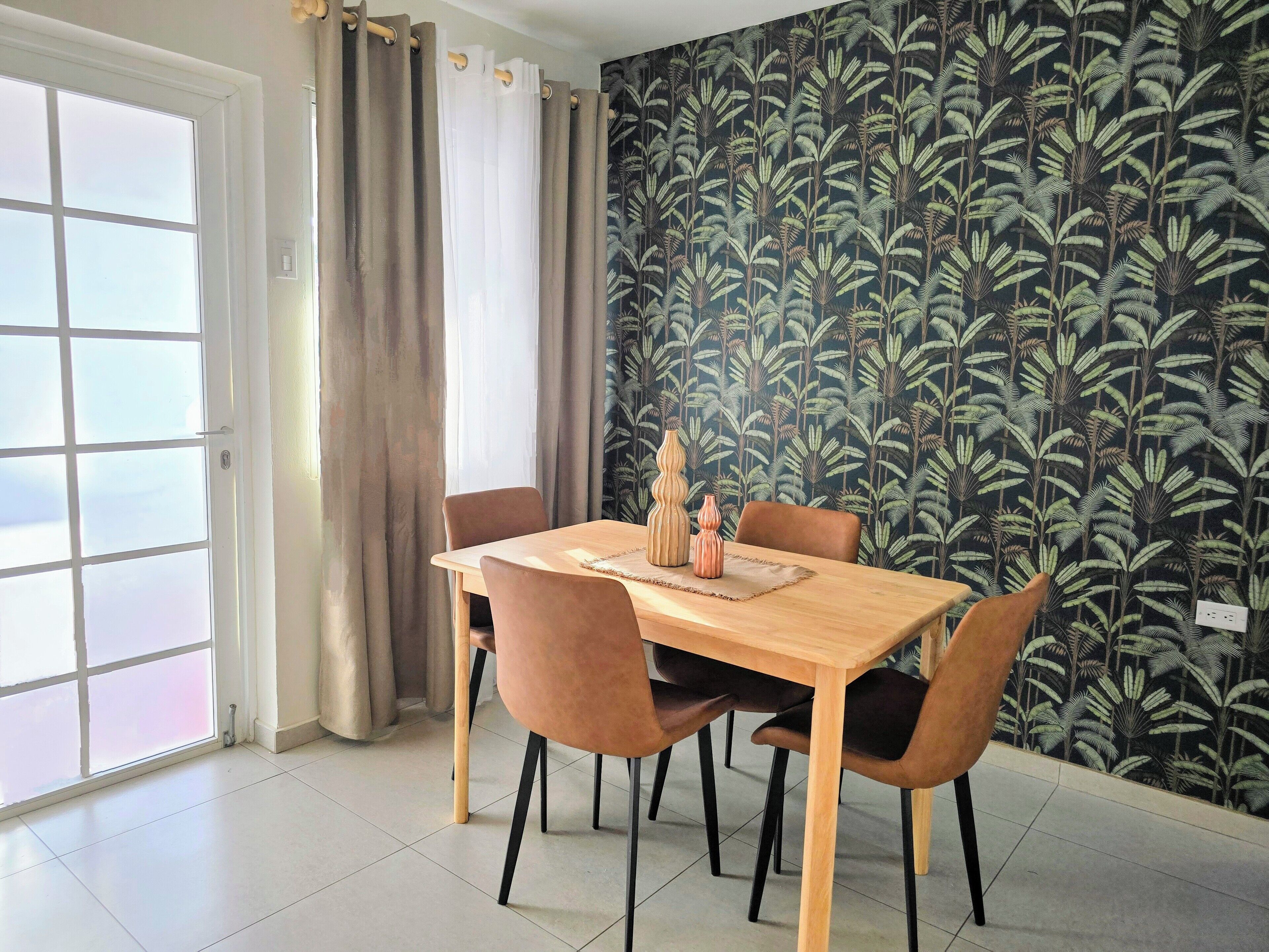 A dining area with a table and chairs, a window with curtains, and a wall with a leafy pattern.