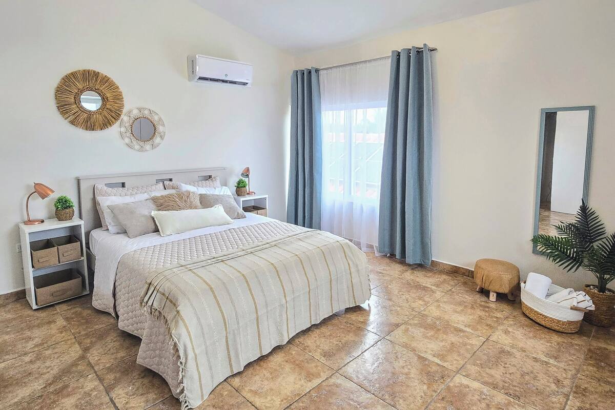 A bedroom with a bed, nightstands, a ceiling fan, an air conditioner, a window with curtains, and a mirror.