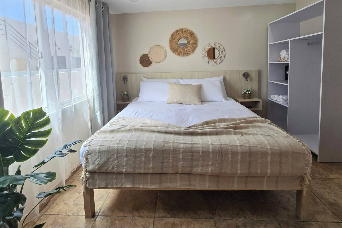 A neatly made bed with a beige blanket, a bedside table with a lamp, and a wall with decorative mirrors and wall art.