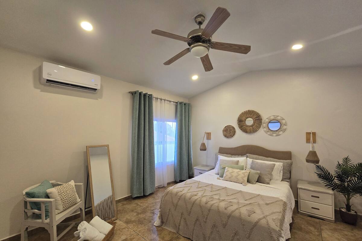 A bedroom with a bed, a ceiling fan, an air conditioning unit, a bedside table, a nightstand, a mirror, a window with curtains, and a potted plant.