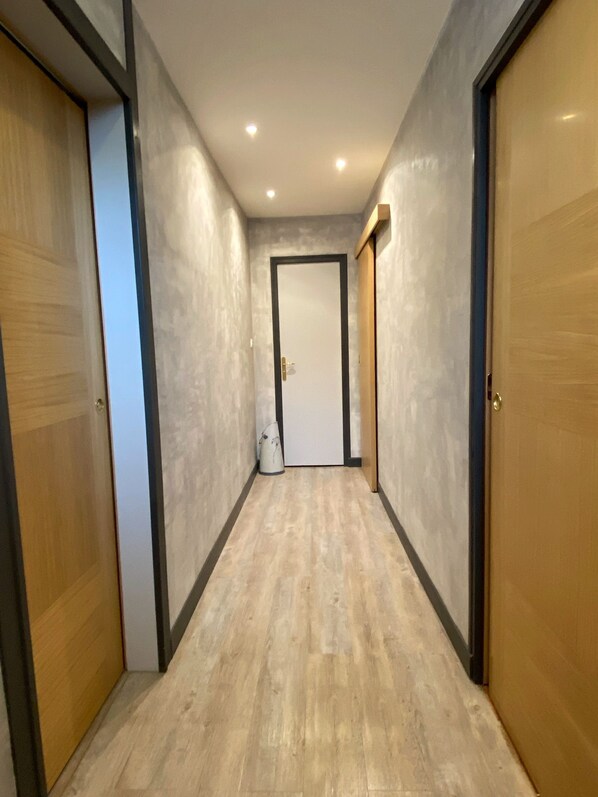 Un pasillo con puertas de madera, una puerta blanca al final y un suelo de color claro.