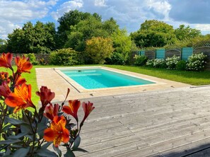 A rectangular swimming pool with a wooden deck in front, surrounded by lush greenery and a brick wall.