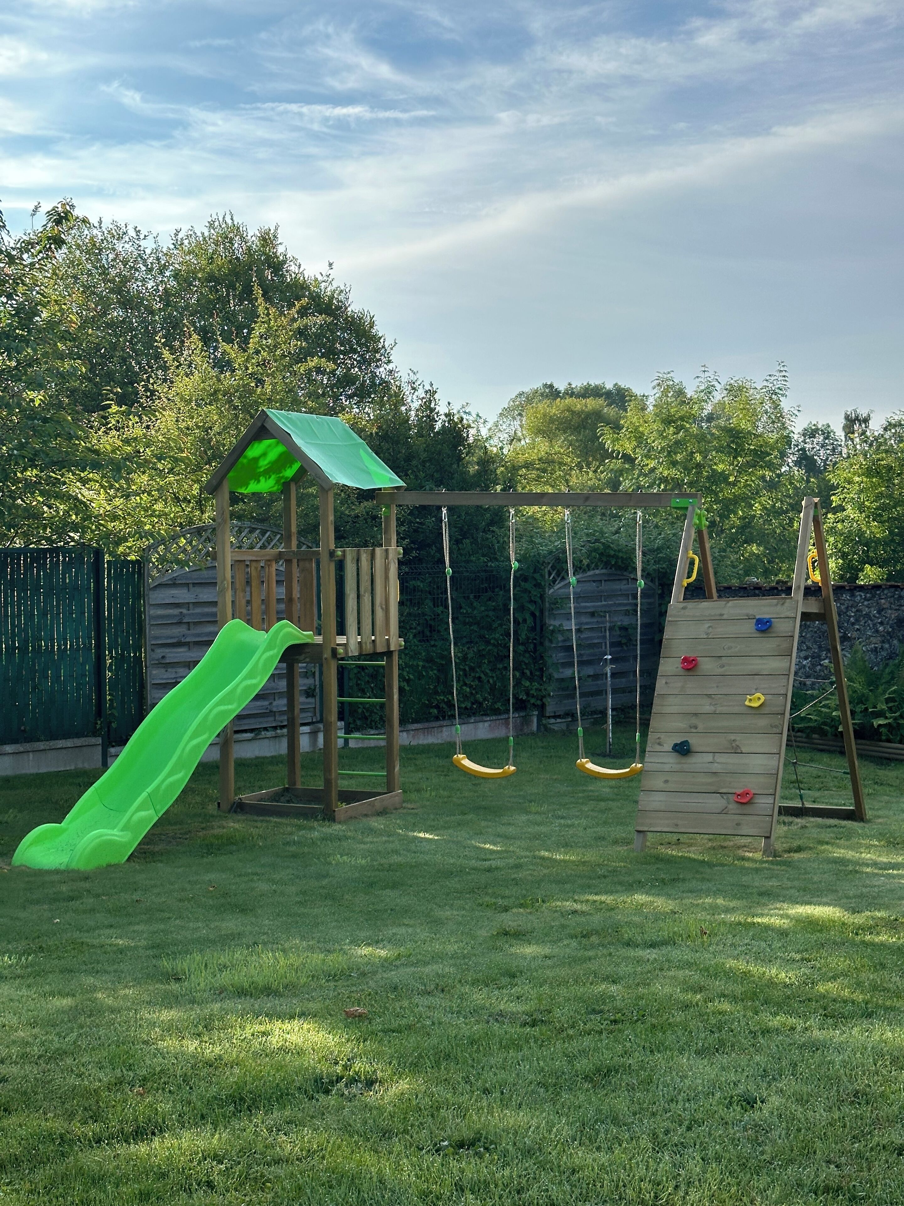 A wooden play structure with a slide and climbing wall, surrounded by a grassy yard and trees.