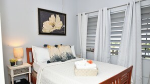 Una cama bien tendida con una colcha blanca y una almohada decorativa. Hay una mesita de noche con una lámpara y una planta. Un cuadro enmarcado cuelga de la pared sobre la cama.