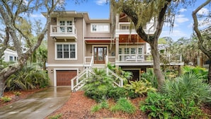 A two-story house with a front porch, a garage, and a well-maintained garden with various plants and trees. - Beautiful 2nd Row Beach Home w/ Beautiful Views (Hilton Head Island)