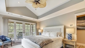 A bedroom with a large bed, two bedside tables with lamps, a ceiling fan, and a view through large windows. - Beautiful 2nd Row Beach Home w/ Beautiful Views (Hilton Head Island)