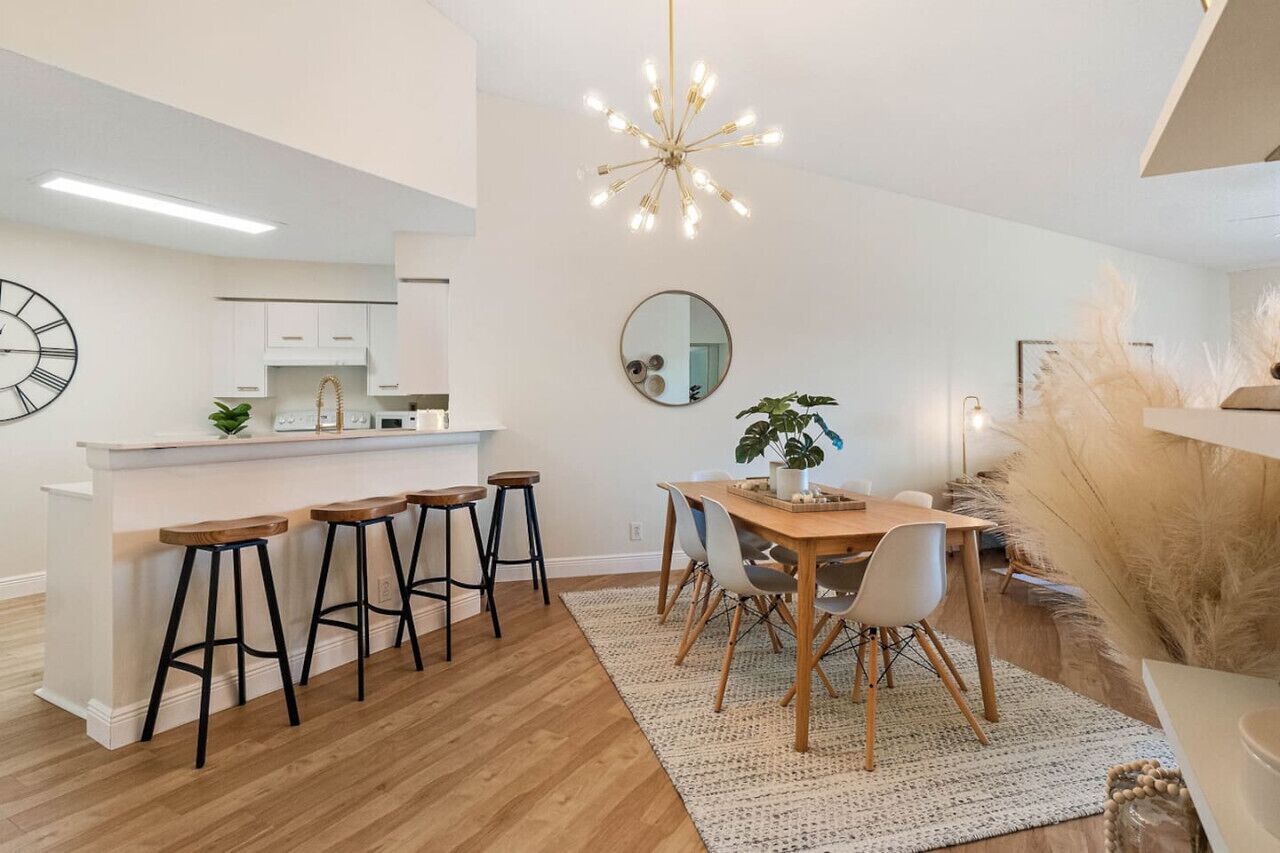 A modern kitchen with white cabinets, a bar area with stools, a dining table with chairs, and a decorative mirror.