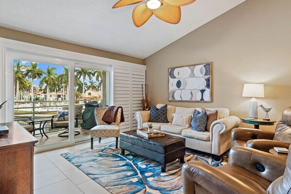 A spacious living room with a large window, a ceiling fan, a sofa set, a coffee table, and a decorative lamp. - Gorgeous Updated 2 Bedroom Lake View Unit in (Isla Del Sol)