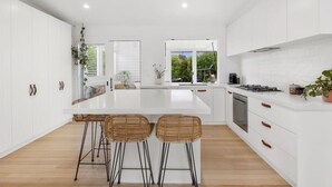 A modern kitchen with white cabinetry, a central island with stools, a dishwasher, and a window with a view of greenery. - Breezy Beach Vibes 350m from Shore (Torquay)