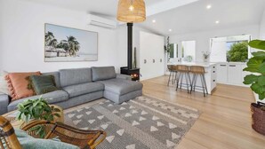 A modern living room with a grey sofa, a patterned rug, a wicker chair, a hanging light fixture, and a kitchen area with white cabinets and a bar stool. - Breezy Beach Vibes 350m from Shore (Torquay)