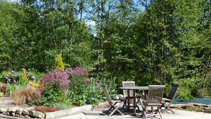 An outdoor patio area with a concrete table and four chairs, surrounded by lush greenery and flowering plants.