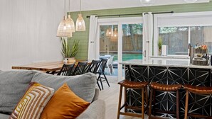 A modern kitchen with a dining area, featuring a bar counter, pendant lights, and a view of the backyard.