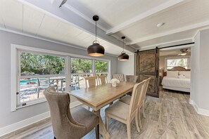 A dining area with a large table, chairs, and pendant lights. There is a view of a pool and greenery outside.
