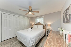 A bedroom with a bed, a ceiling fan, a nightstand with a lamp, a window with blinds, a wardrobe, and a wall with a framed picture.
