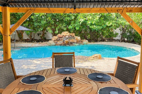 A wooden deck with a pool table and chairs, surrounded by a pool and a rock waterfall.