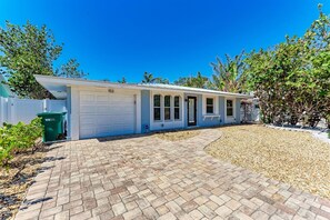 A single-story house with a white garage door, a brick driveway, and a small garden area with gravel and plants. - La Cocolia located on north end of Ami. Lagoon Pool, Walk to Pine Ave. (Anna Maria)