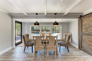 A dining area with a wooden table and chairs, a sliding barn door, and a view of a swimming pool through large windows. - La Cocolia located on north end of Ami. Lagoon Pool, Walk to Pine Ave. (Anna Maria)