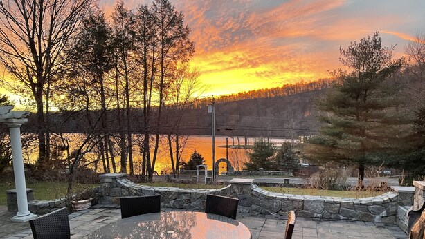 Une terrasse avec une table ronde et des chaises, donnant sur une étendue d’eau et une zone boisée, au moment du coucher du soleil.