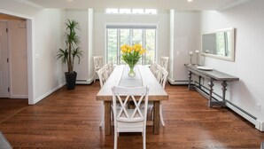 Une salle à manger avec une longue table en bois, des chaises blanches, une plante en pot et un vase rempli de fleurs jaunes.