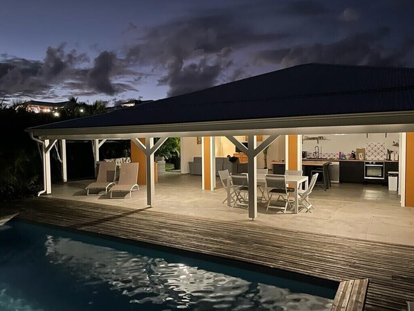 A covered outdoor area with a pool, lounge chairs, and a kitchen area.