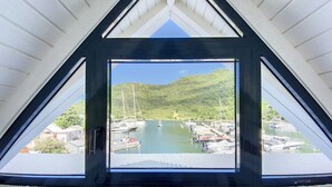A view from a room with a triangular window overlooking a marina with boats and a hilly landscape. - Marina Beach 1, entre la plage et la marina (Anse Marcel)