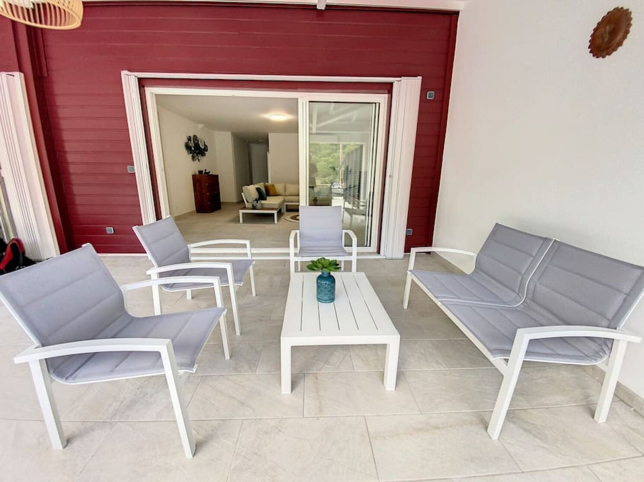A patio with white furniture, including two sofas and two armchairs, a small table with a vase, and a sliding glass door leading to an interior space with a sofa and a coffee table.
