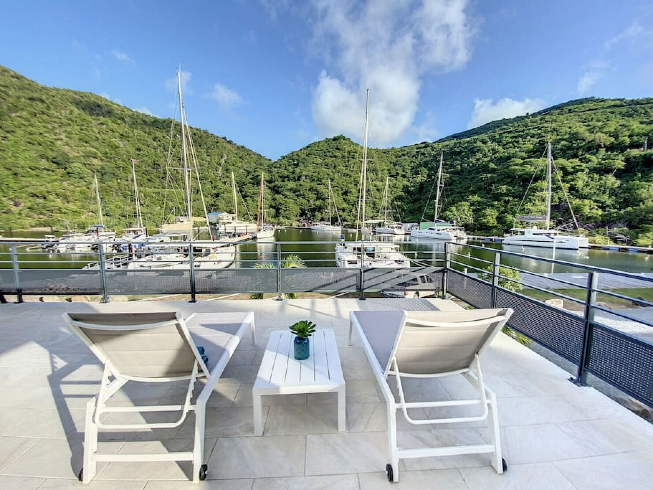 Une terrasse avec deux fauteuils blancs et une petite table, donnant sur une marina où sont amarrés plusieurs voiliers.