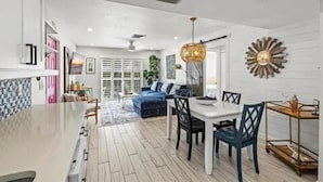A modern kitchen with a central island, a dining area with a white table and blue chairs, and a living space with a blue sofa and a large window. - Modern Beach House, 1 block from beach! (Holmes Beach)
