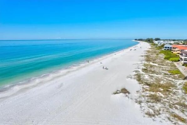Una playa con agua turquesa cristalina, arena blanca y algunos edificios a la derecha.