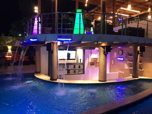 A pool area with a bar and lounge seating, surrounded by a pool deck and a covered seating area with a view of the pool.