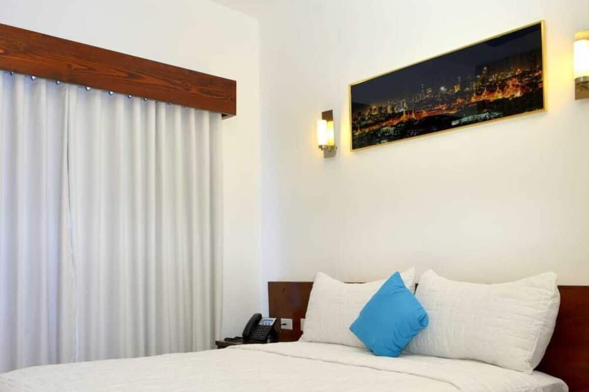 Una habitación de hotel con cama, ventilador de techo, un cuadro de un paisaje urbano nocturno y un teléfono en la mesita de noche.