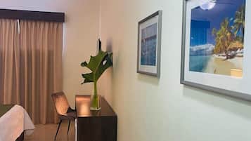 A hotel room with a bed, a desk with a plant, a chair, and framed pictures on the wall.