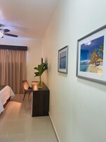 A hotel room with a bed, a desk with a plant, a chair, and framed pictures on the wall.