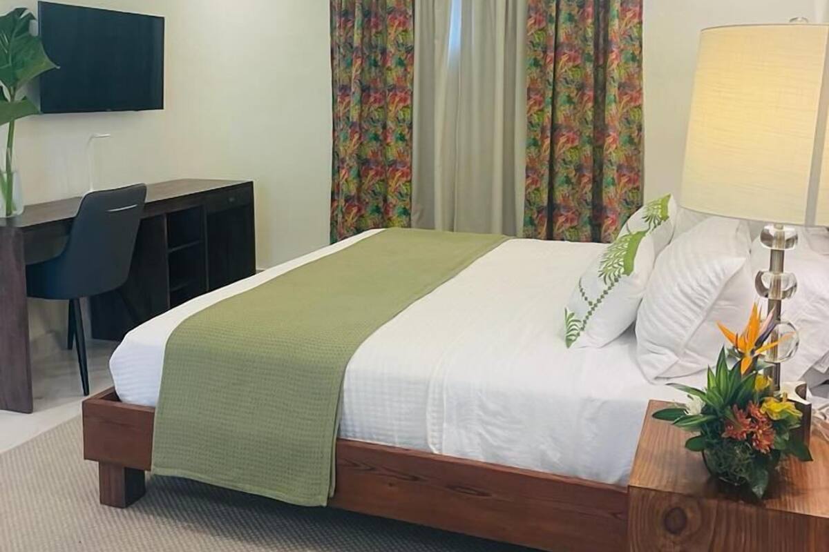 Habitación de hotel con cama, escritorio, silla, televisor y mesita de noche con lámpara y flores.