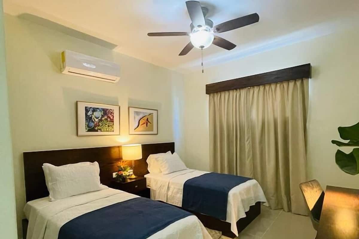 Habitación de hotel con dos camas, ventilador de techo, aire acondicionado, escritorio y una ventana grande con cortinas.