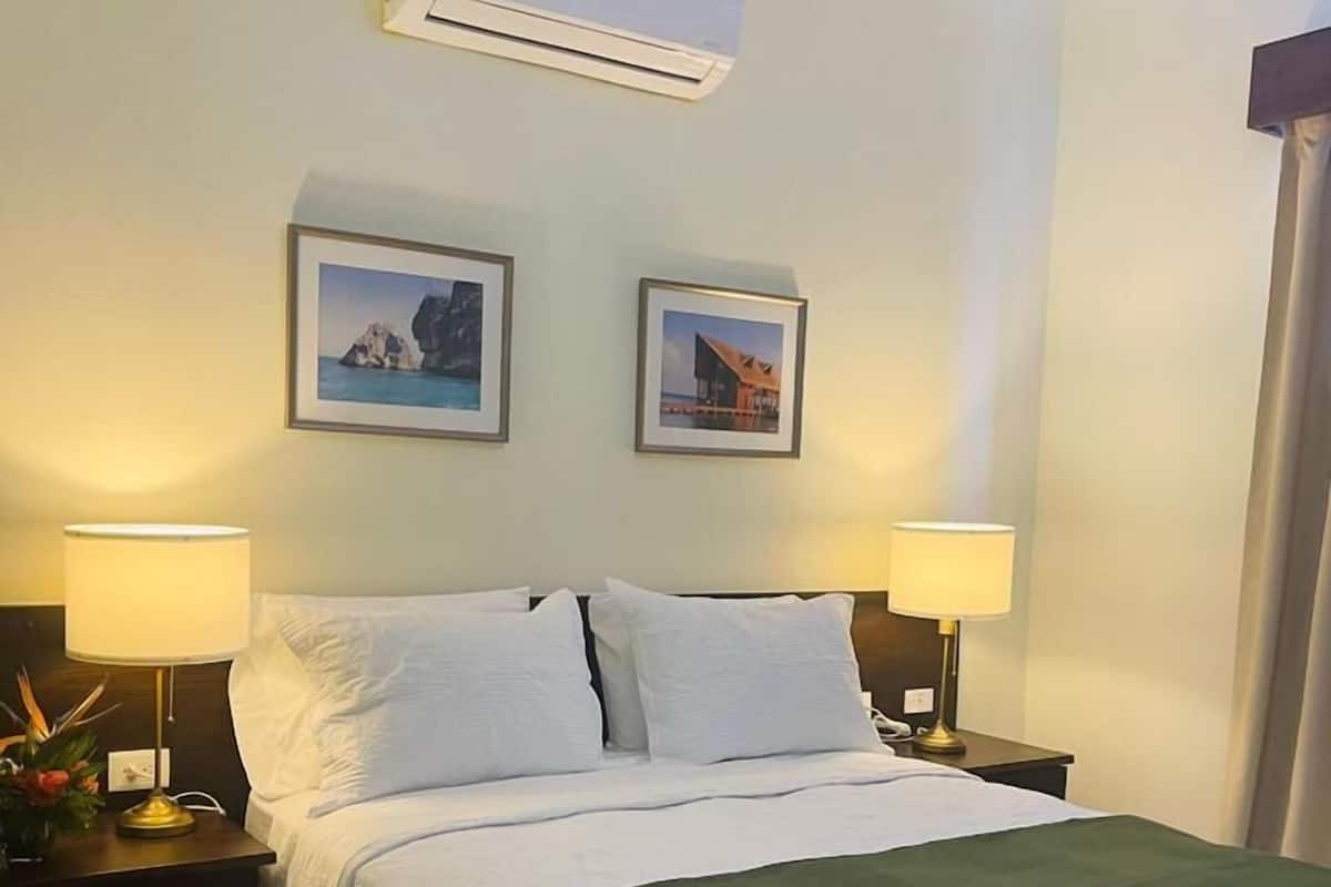 Habitación de hotel con cama, dos lámparas de noche, un equipo de aire acondicionado y dos cuadros en la pared.
