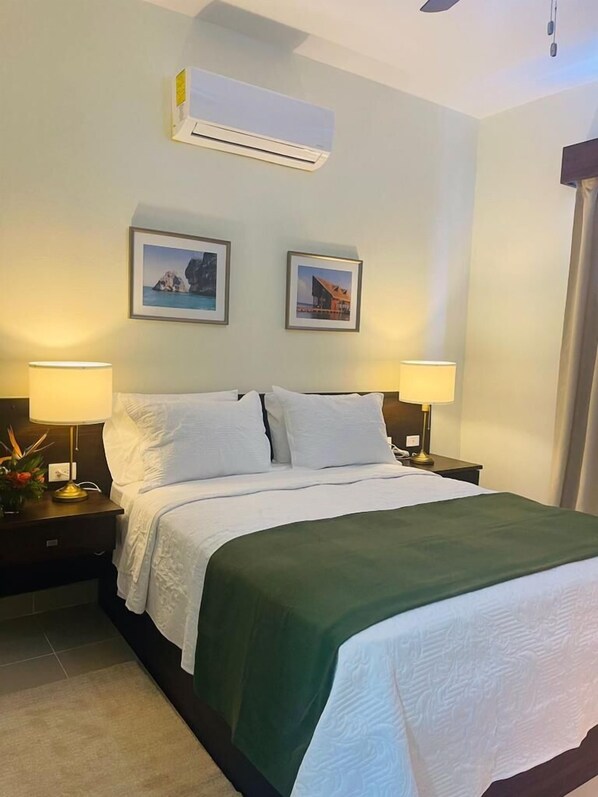 A hotel room with a bed, two bedside lamps, an air conditioning unit, and two framed pictures on the wall.