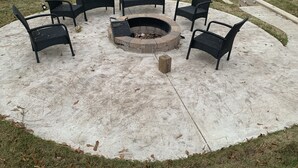 Un espacio de estar circular al aire libre con seis sillas de mimbre negro y un brasero en el centro.