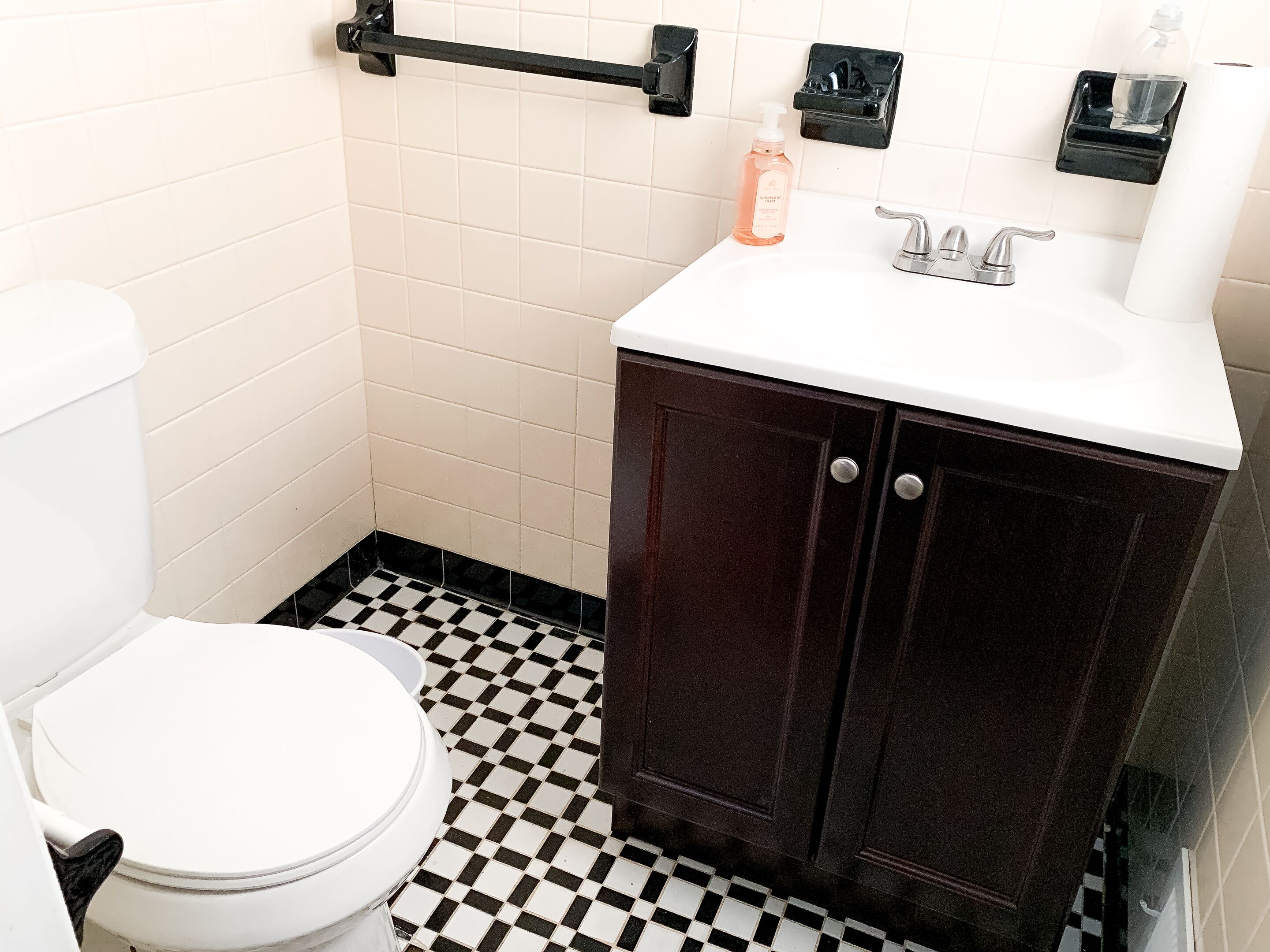 Une salle de bain avec des toilettes blanches, un carrelage noir et blanc, un meuble en bois foncé avec un plan de travail blanc et un porte-serviettes noir.