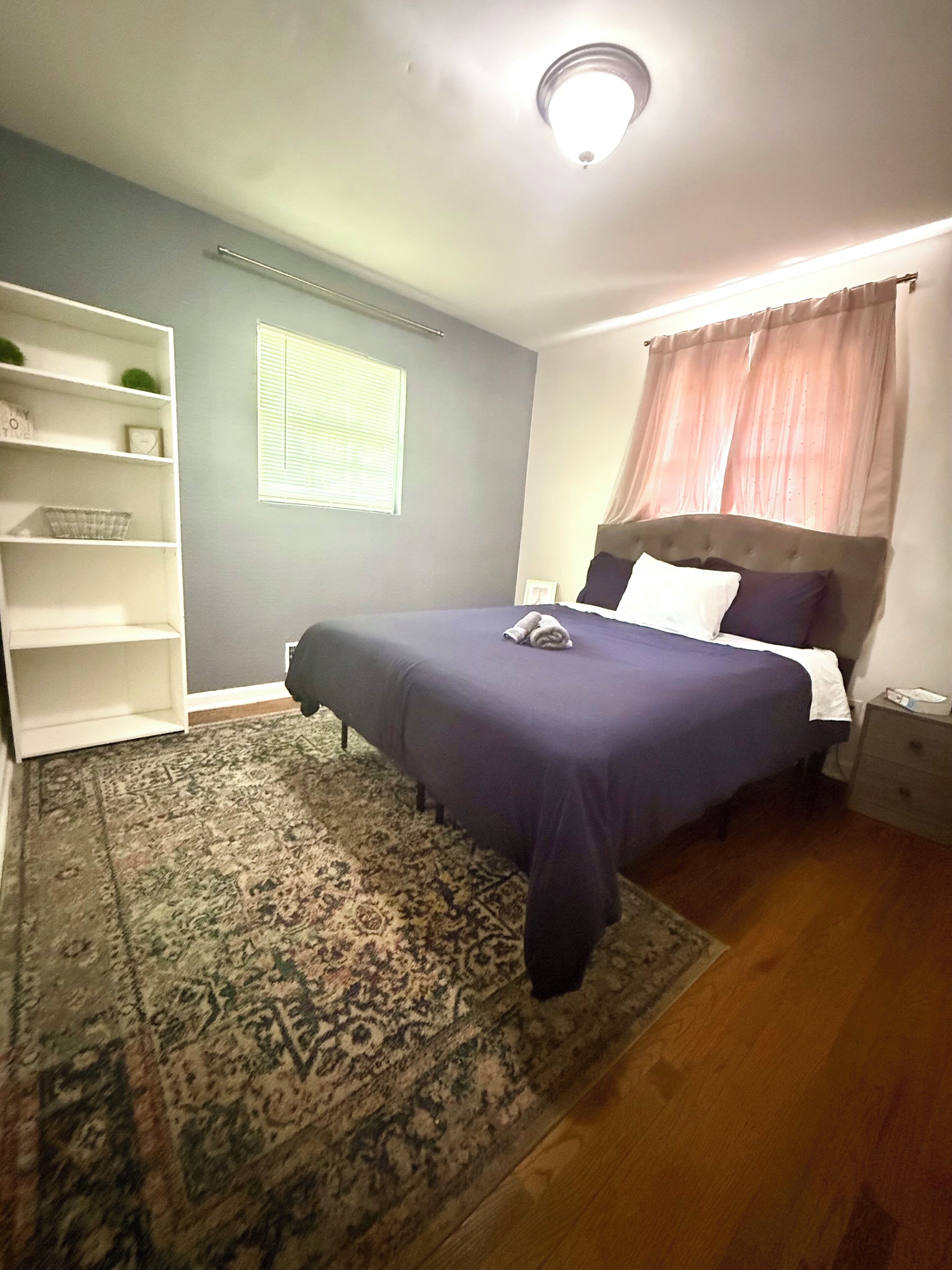 Une chambre à coucher comprenant un lit, une table de chevet, une étagère, une fenêtre avec des stores et un tapis à motifs.