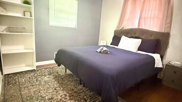 Une chambre à coucher comprenant un lit, une table de chevet, une étagère, une fenêtre avec des stores et un tapis à motifs.