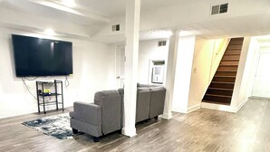 Una sala de estar con televisor de pantalla plana, sofás grises y una escalera.